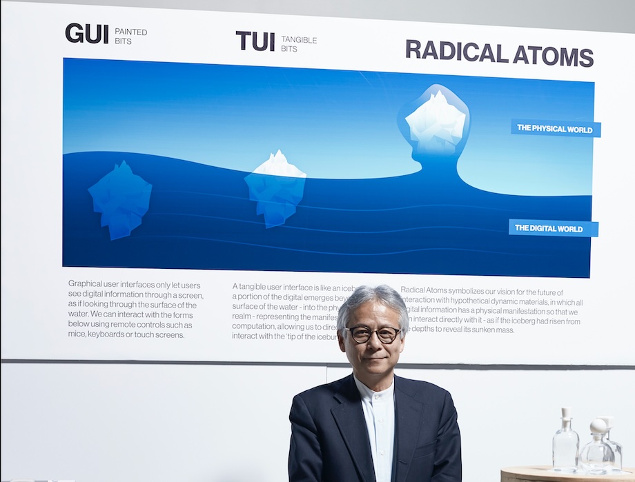 Portrait of Hiroshi Ishii in front of a diagram showing GUI, TUI, and Radical Atoms as icebergs of increasing size rising from the digital world into the physical world. Beside him on a table are glass bottles from his Music Bottles project, an early tangible interface that let people control music by opening and closing physical bottles.